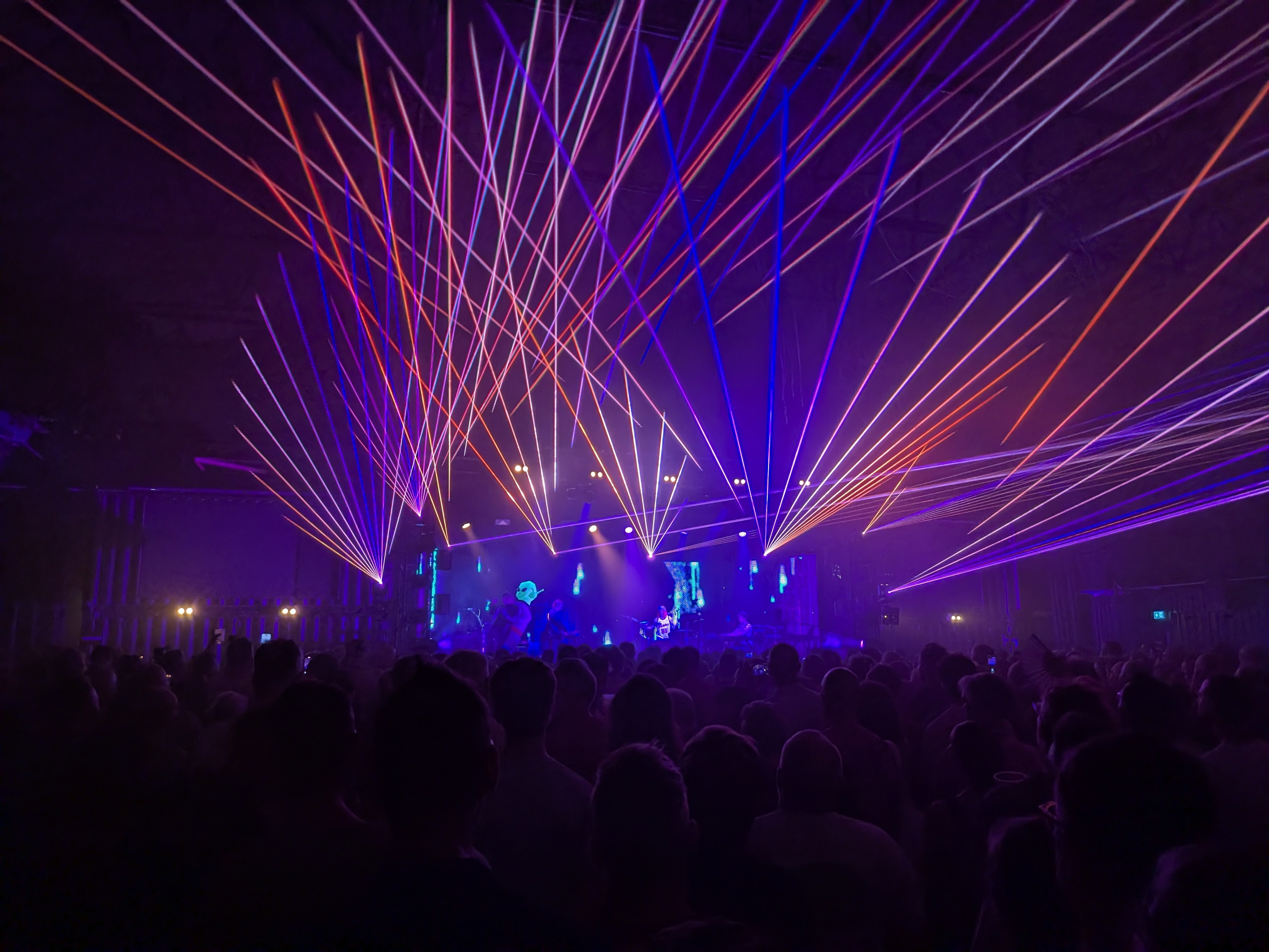 Photo at a gig over a large crows.  The stage is illuminated in dark blue.  Red, purple and blue lasers span the ceiling.