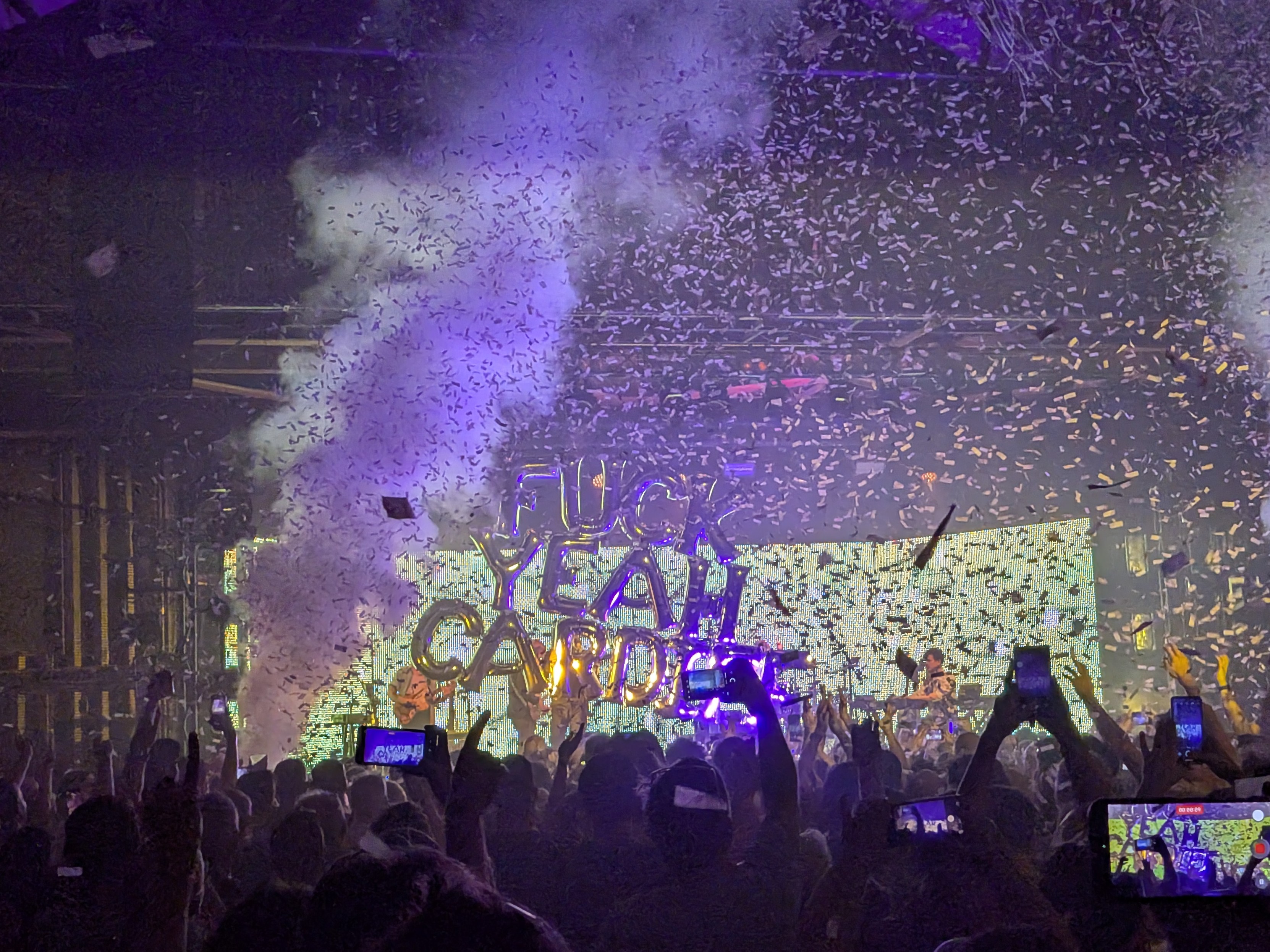 A photo at a gig.  A bright green background screen is a backdrop to a large silver balloon, approximately 20 feet tall, in lettering reading "FUCK YEAH CARDIFF".  A smoke machine blasts from bottom left of frame.  Crowd cheers and records on phone as masses of small paper pieces fall from the ceiling.