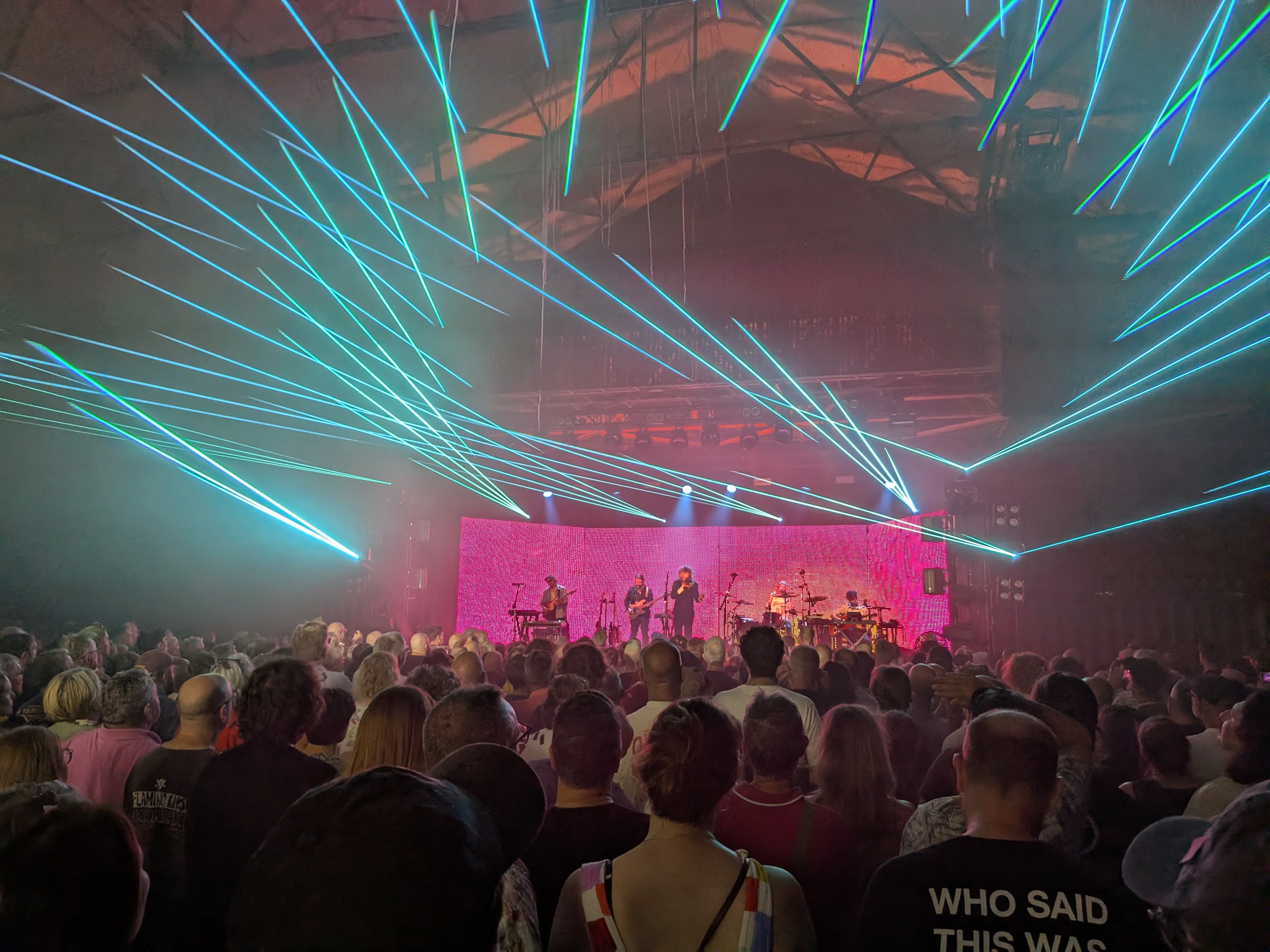 Photo at a gig.  The band on stage is framed by magenta light.  Sky blue lasers span the ceiling.